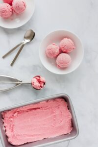 A tub of strawberry gelato with several ice cream scoops in a bowl.
